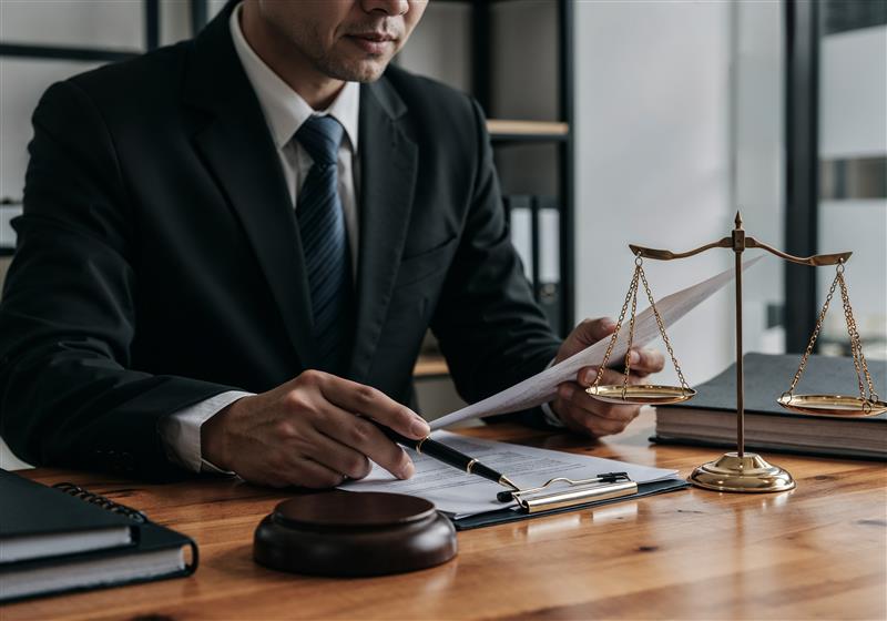 A lawyer in a black suit reviewing documents at a wooden desk, with a gavel, legal scale, and clipboard in front of him in an office setting.