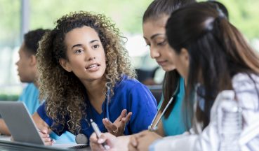 Three medical trainees sit in their classroom in front of a laptop and work on an assignment together.  One young student talks as the others listen and take notes.