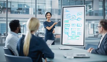 Female Operations Manager Holds Meeting Presentation for a Team of Economists. Asian Woman Uses Digital Whiteboard with Company Project Management Plan, Charts, Data. People Work in Business Office.