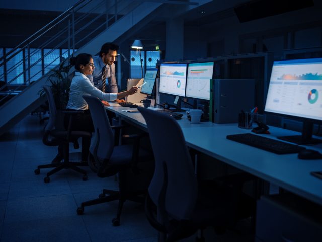 Late at Night In Modern Office: Businessman and Businesswoman Work on Desktop Computer, Having Discussion, Finding Problem Solution, Finishing Project. Successful Dedicated Responsible Office Workers